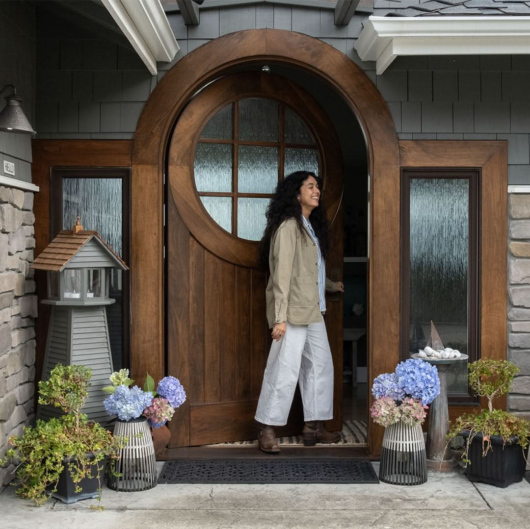 A woman smiling as she steps out of a home through a round-top wooden front door with glass panels. The door is framed by matching sidelights, surrounded by stone and shingle siding, and decorated with hydrangeas and potted plants.