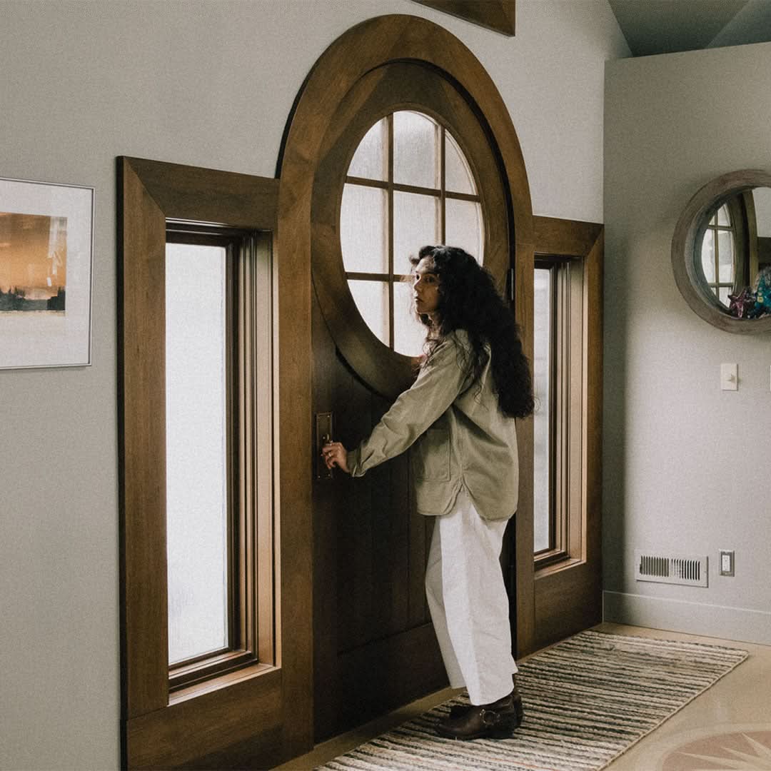 An interior view of a round-top wooden front door with glass panels, framed by sidelights, in a cozy entryway. A woman stands near the door, reaching for the handle, with soft natural light illuminating the space