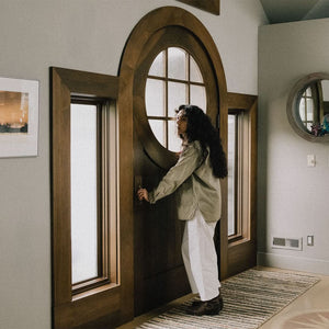An interior view of a round-top wooden front door with glass panels, framed by sidelights, in a cozy entryway. A woman stands near the door, reaching for the handle, with soft natural light illuminating the space