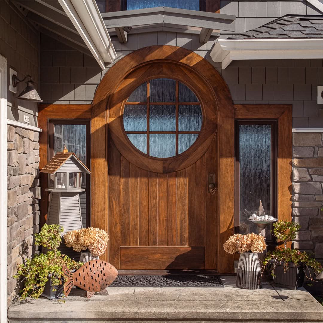 A close-up exterior shot of a round-top wooden front door with a circular glass window and matching sidelights. The doorway is accented with stone walls, decorative plants, and a charming lighthouse-style ornament.