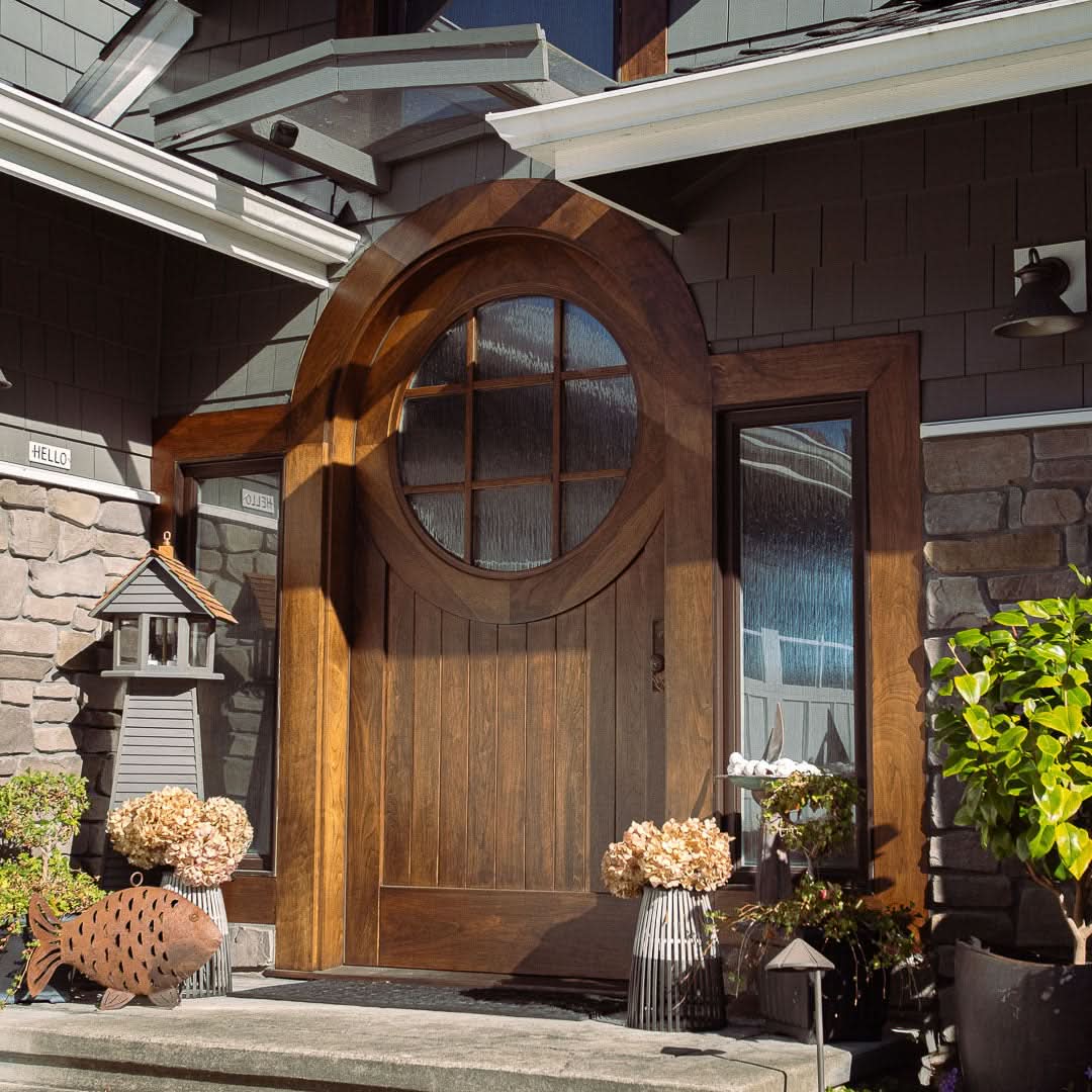 A side-angle view of a round-top wooden front door with glass panels and sidelights, surrounded by stone and shingle siding. The entrance features decorative plants, a fish-shaped ornament, and a welcoming atmosphere in natural sunlight.