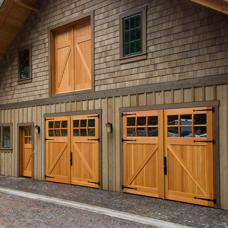 Row of solid wood carriage doors with Z-brace detailing and divided-lite glass panels, set into a shingle-clad barn-style building with matching trim and vertical siding.