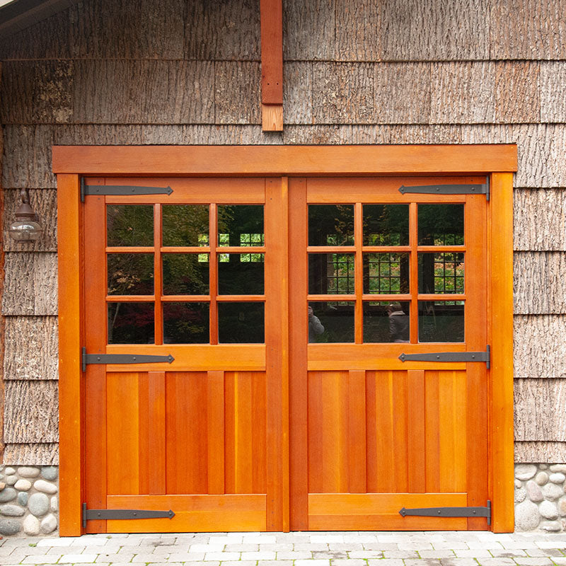 Natural wood carriage garage doors with divided glass windows and black strap hinges, installed on a shingle-sided garage exterior in daylight.