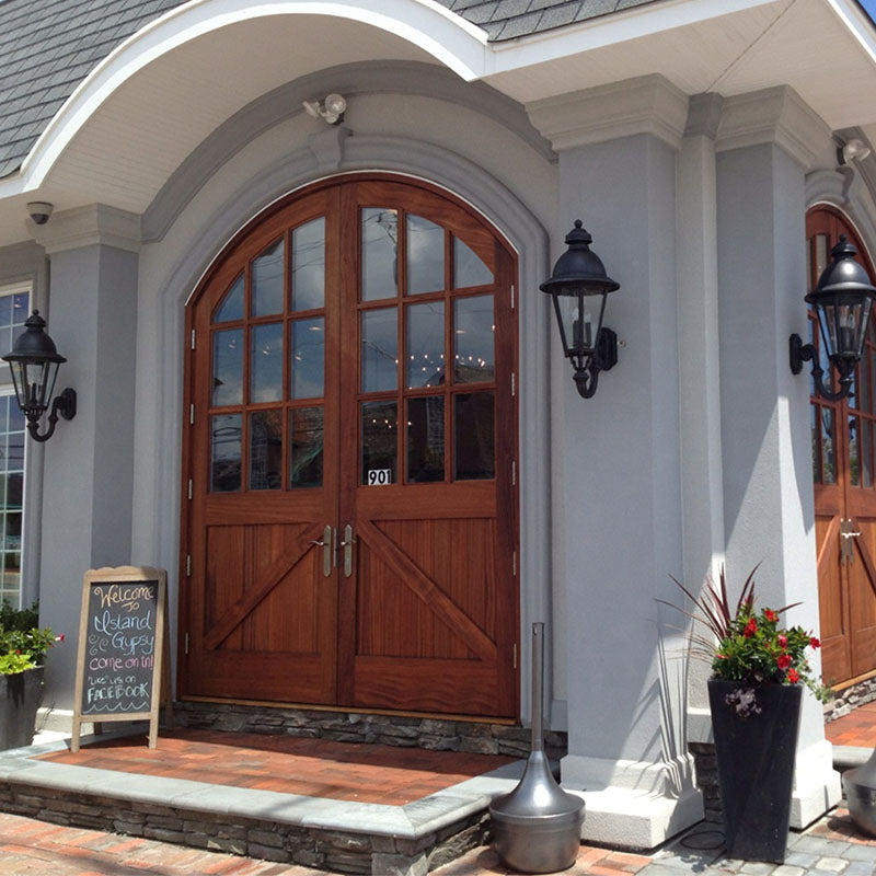 Arched wooden carriage-style double doors with glass panes, installed at the entrance of a light gray stucco building with black lantern sconces, brick walkway, and potted plants framing the doorway.