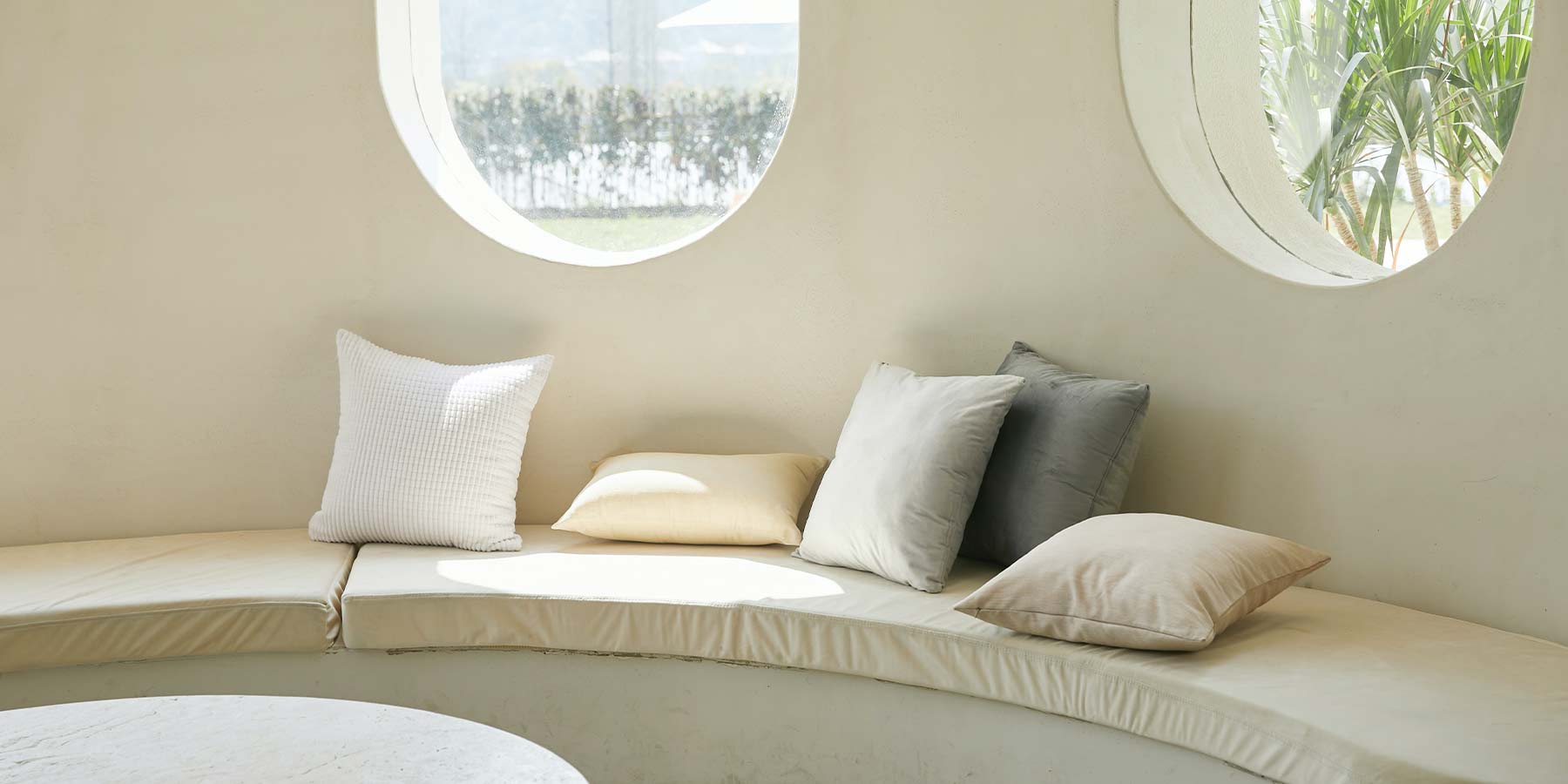 Beach House interior. Shoot of a circular corner white bench. There ae six light colors pillows over the couch and two circular windows above them.
