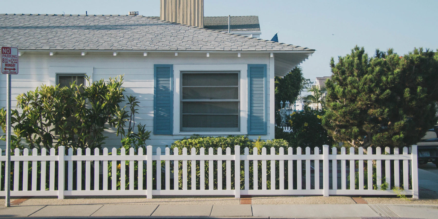 Street curbside corner featuring a white house with a open blue shutters and a white fence.