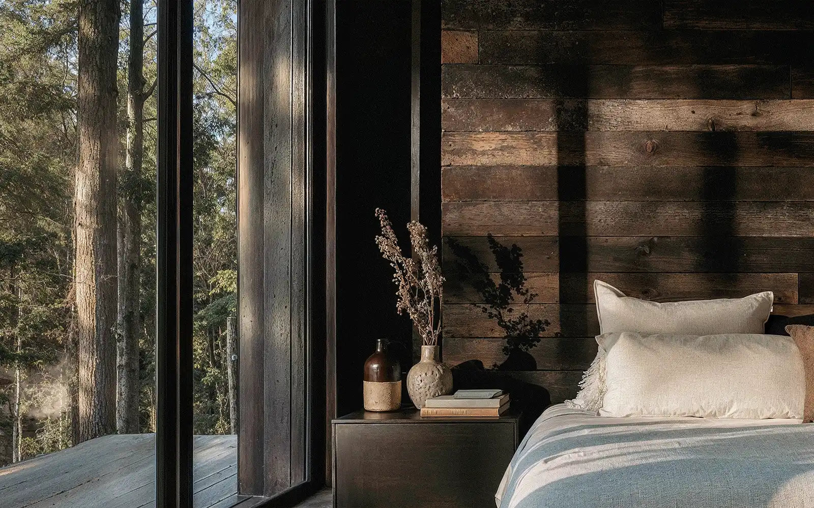 Cozy bedroom with large window showcasing trees outside. Rustic wooden walls, soft white bedding, and a nightstand with dried flowers convey tranquility.