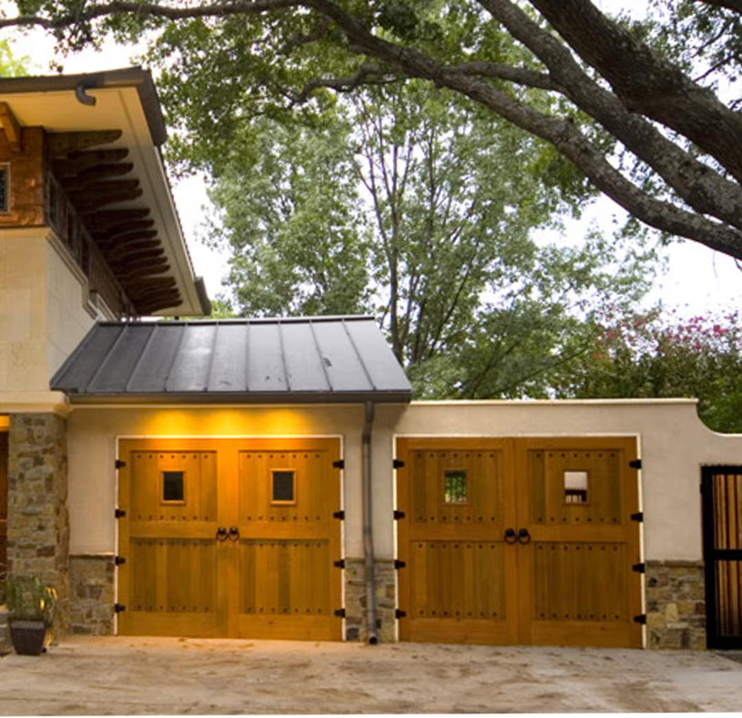 Set of carriage garage doors with a single window panel and rustic ring pull hardware, installed on a residential exterior in daylight.