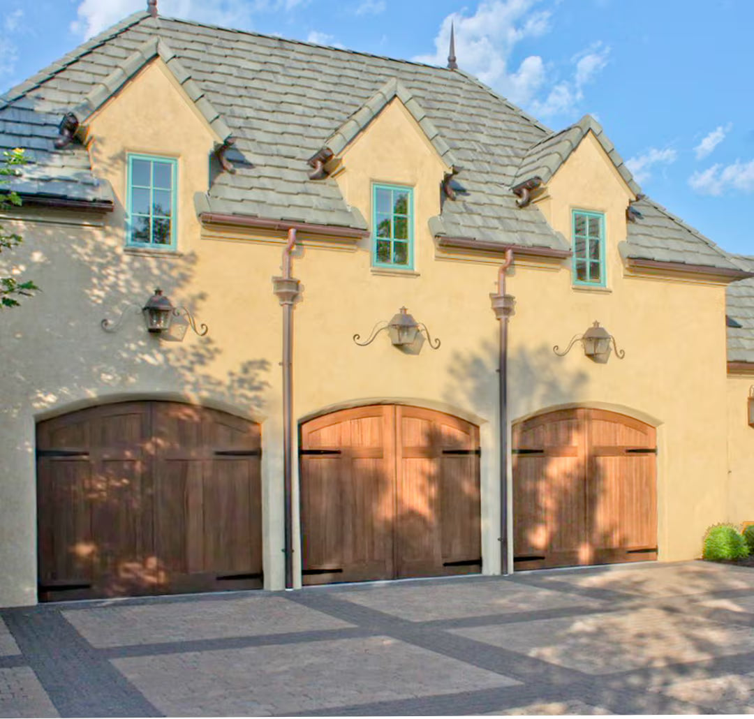 Unfinished arched carriage garage door with strap hinges, shown on a residential exterior in daylight with exposed natural wood tones.