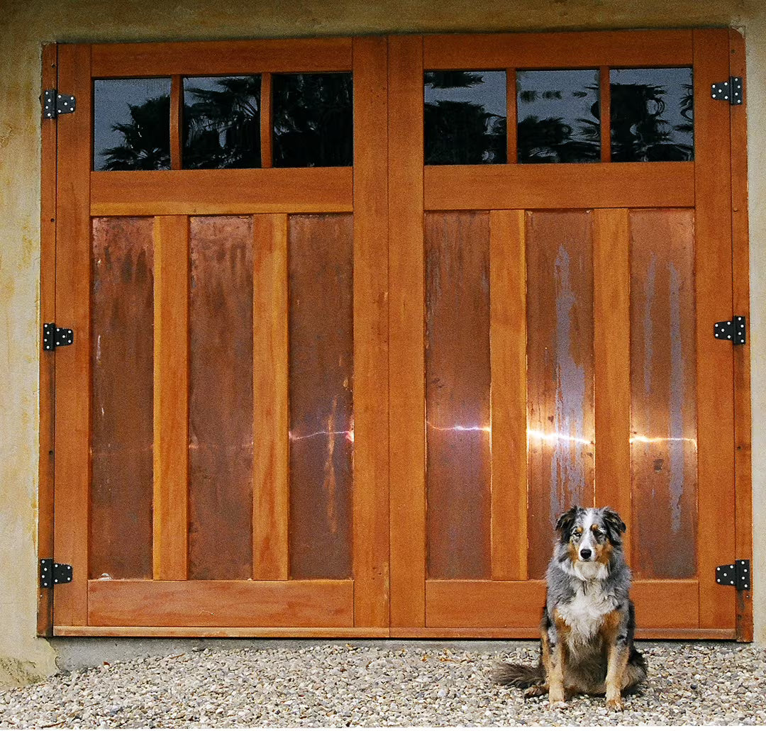 Carriage garage door with a mid-brace design, galvanized metal panels, and small hinges, installed on a residential exterior in daylight.