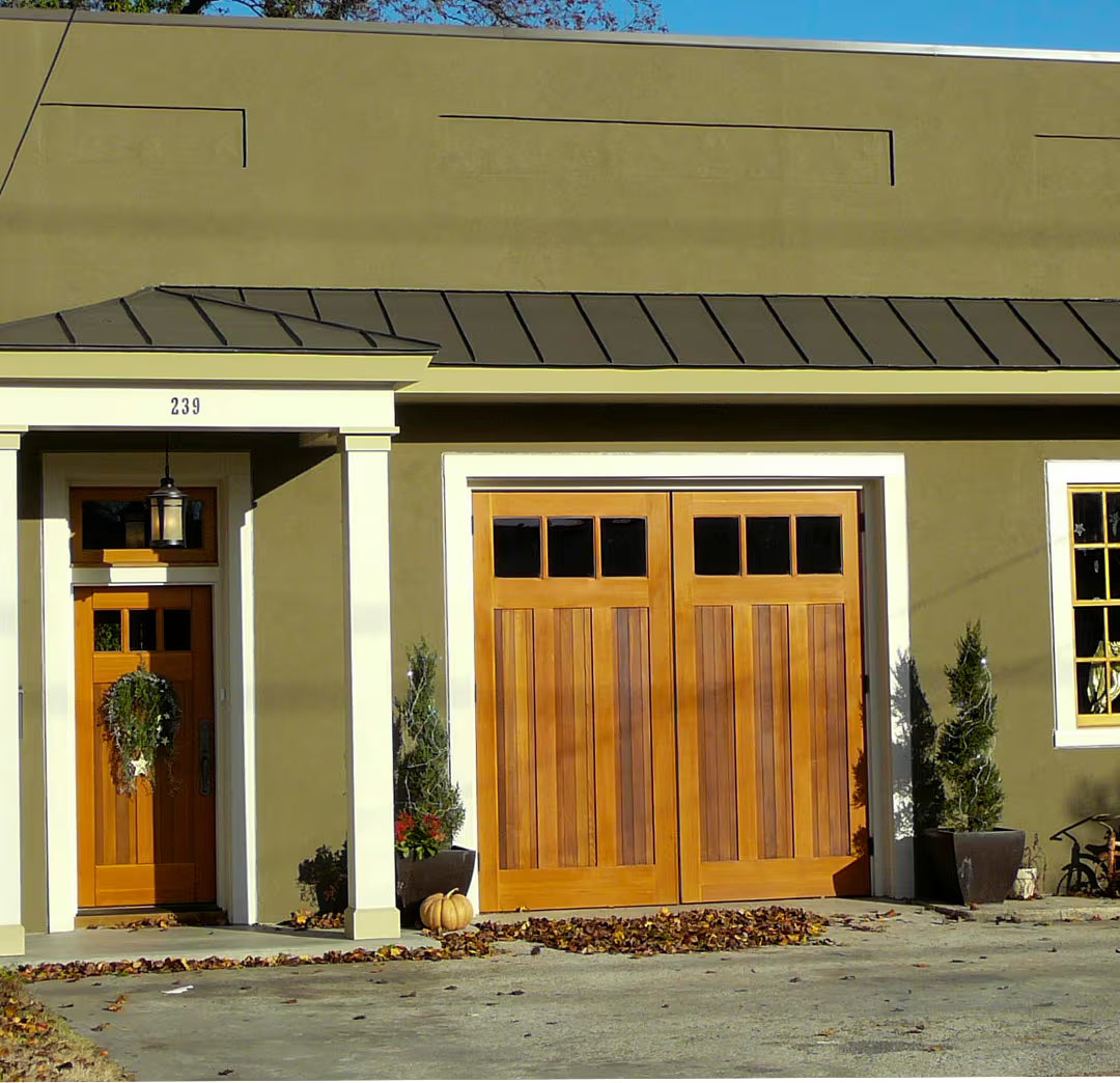 Vertical plank carriage garage door with three clear glass panels, shown alongside a matching wood entry door on a residential exterior in daylight.