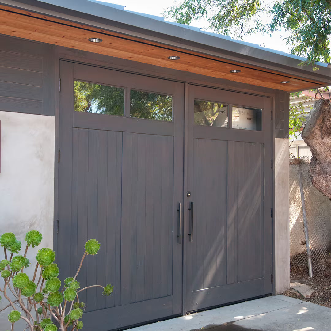 Dark stained vertical plank carriage garage door with two clear glass panels and long metal pull handles, shown on a residential exterior in daylight.