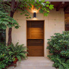 Modern entrance to a building with a wooden door, surrounded by plants and a wall light fixture.