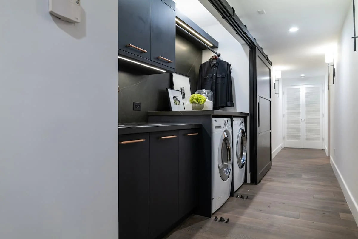 Modern laundry room with sleek black cabinets and copper handles. A washer and dryer sit below a countertop with framed photos and a plant.