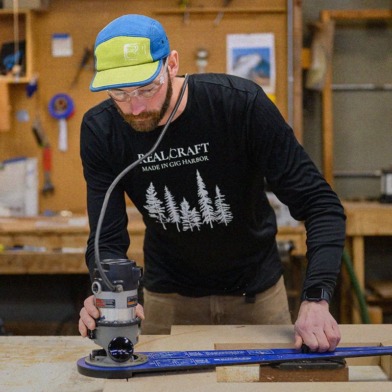A man in a baseball cap and safety glasses uses a router on wood in a workshop. He's focused on his task, creating an atmosphere of concentration.