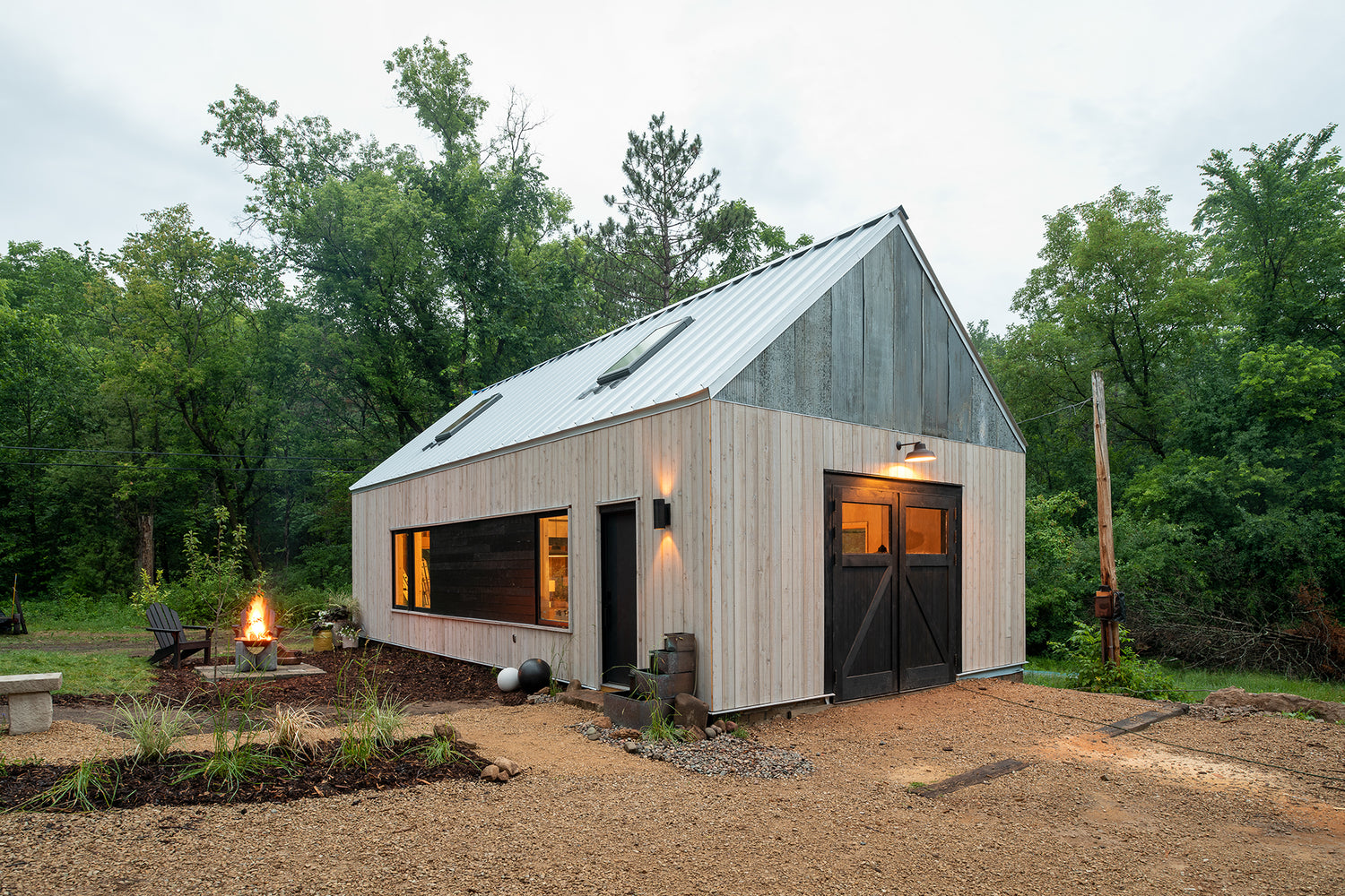 Modern cabin with a steep metal roof and wood siding, situated in a lush forest. A lit fire pit and chairs create a warm, inviting atmosphere.
