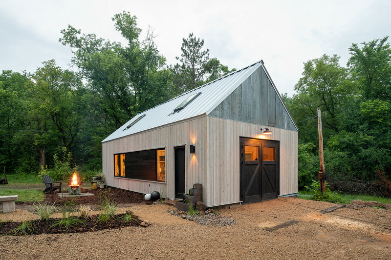 Modern cabin with a steep metal roof and wood siding, situated in a lush forest. A lit fire pit and chairs create a warm, inviting atmosphere.