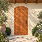 RealCraft Tivoli arched door in Cherry, with six vertical planks and black hardware, in a covered stucco courtyard with patterned stone paving and soft foliage shadows.