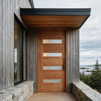 A modern wooden front door with four horizontal frosted glass panels, set in a gray wood-clad house entryway with stone walls and an outdoor view of the ocean and trees under a cloudy sky.