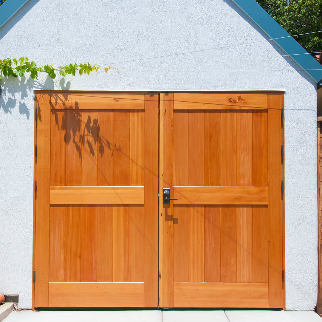 Double solid wood carriage garage doors with a natural finish and flat panel detailing, installed on a white stucco exterior in bright daylight.
